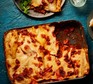 Baking dish of lasagne with a portion removed and put on plate with a green salad